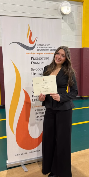 Angelina Vataj holding her Richard Berman Human Rights Leadership Award beside an HHREC banner
